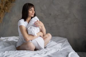 امرأة تقف بهدوء قرب النافذة في ضوء الصباح الخافت، تتأمّل جسدها بعد الولادة.